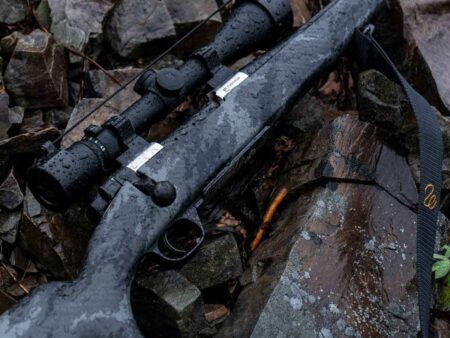 Wet bolt-action rifle with scope resting on rocky terrain in rain