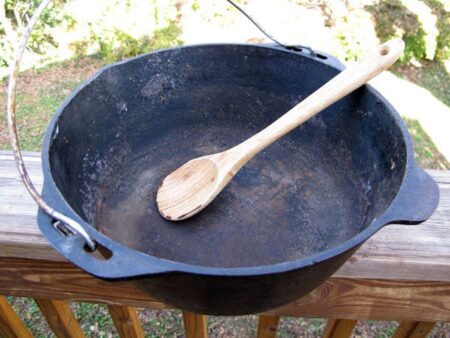 Large seasoned cast iron cauldron with wooden spoon resting on outdoor wooden railing