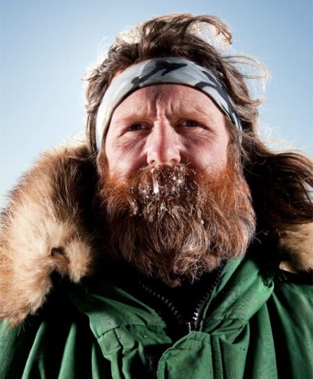 Bearded man with frost-covered beard wearing fur-lined green parka in snowy conditions