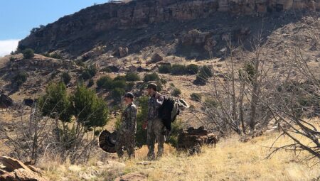 Two hunters in camouflage carrying harvested wild turkey in arid desert landscape.