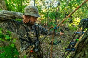 Hunter in camouflage drawing compound bow while standing in forest tree stand