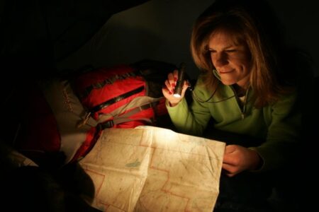 Woman reading map by flashlight in dark tent with red backpack nearby