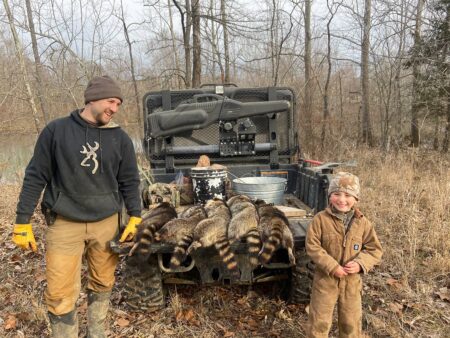 Adult and child display raccoon hunt results beside ATV in wooded area.