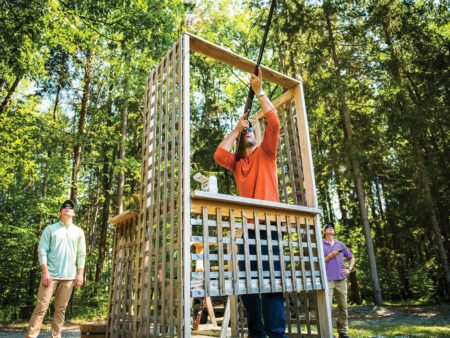 Man shooting shotgun upward from wooden tower while two others watch outdoors.