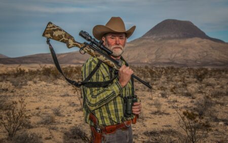 The 7 Essential Traits of a Great Hunting Rifle Man in cowboy hat carrying rifle and binoculars in desert landscape.