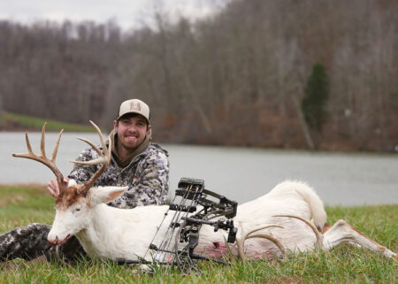 Hunter posing with harvested albino white-tailed deer buck and compound bow outdoors.