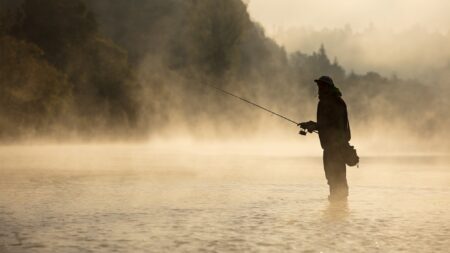 Silhouette of angler fly fishing in misty river at golden sunrise
