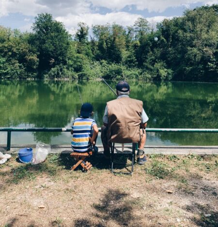 An elderly man and young boy fishing together by a green lake.
