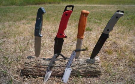 Five outdoor survival knives displayed upright on a wooden log in grass