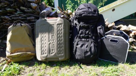 Four travel bags including backpacks and hard shell luggage lined up outdoors near firewood.