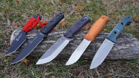 Four fixed-blade survival knives displayed on a log outdoors in grass.