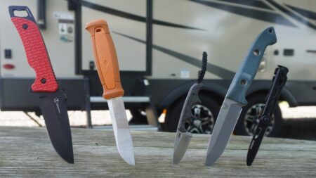 Four outdoor camping knives displayed on wooden surface with RV in background.