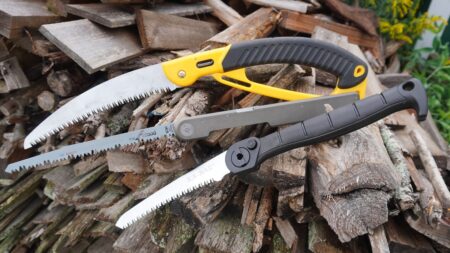Three folding hand saws displayed on a pile of cut firewood outdoors.