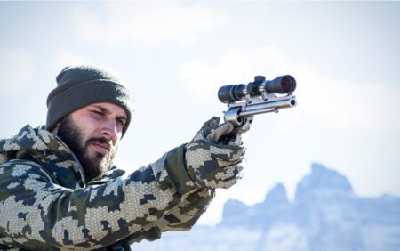 Bearded hunter in camouflage aiming scoped pistol outdoors in snowy mountain landscape