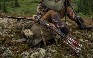 Bowhunter in camouflage gear sitting outdoors with recurve bow and arrows on tundra