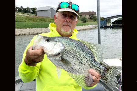 Man in yellow hoodie proudly holds large crappie fish near lake dock.