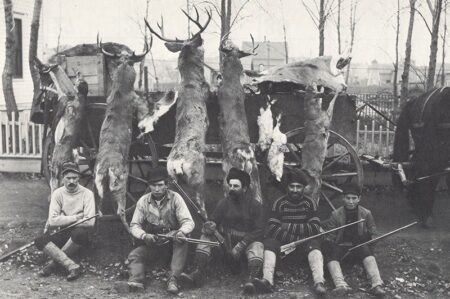 Five hunters posing with rifles and hunted deer carcasses hung on wagon wheels.