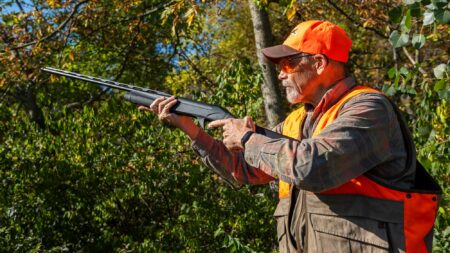Why the Lightweight Shotgun Craze Is, Well, Kind of Crazy Hunter in orange safety vest aiming shotgun in autumn woodland setting