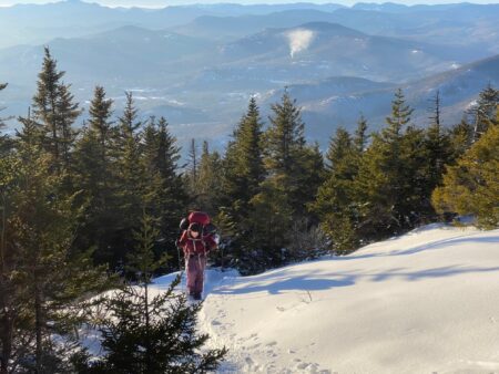 Hiker with red backpack ascending snowy mountain trail surrounded by pine trees
