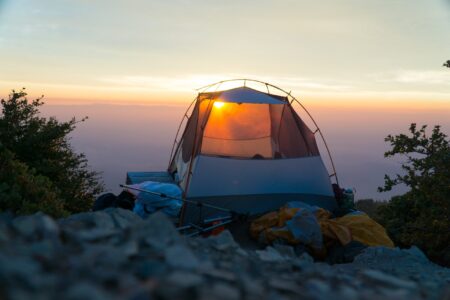 Backpacking tent glowing at sunset on rocky mountain summit with panoramic views.