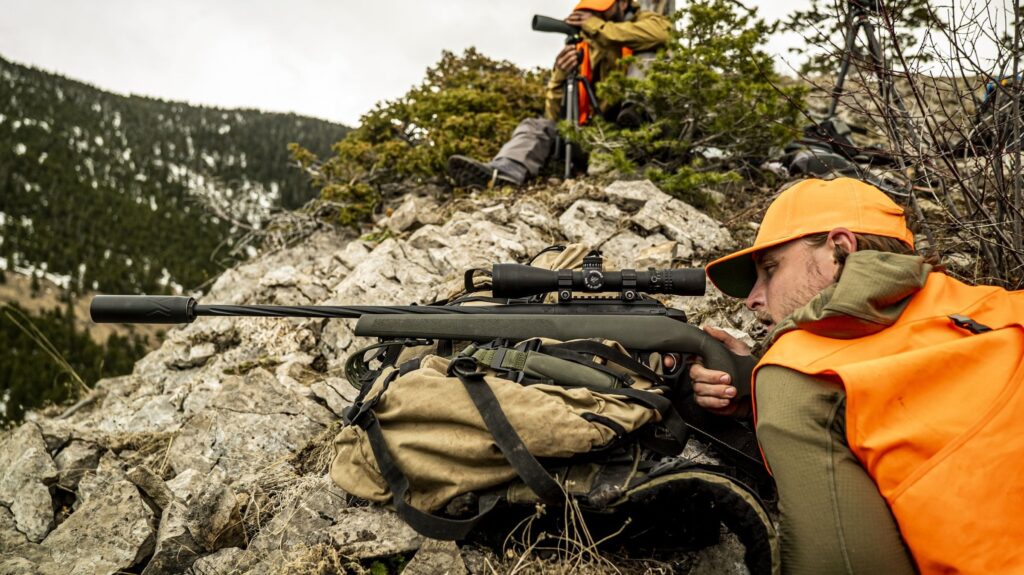 The Best Long Range Rifles, According to Our Testing Hunter in orange vest aims scoped rifle from rocky mountainside while spotter watches nearby.