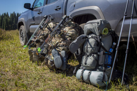 Bowhunting gear with compound bows and backpacks leaning against pickup truck outdoors.