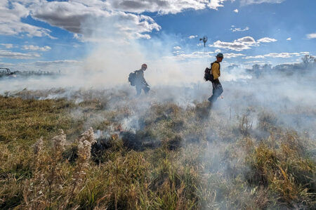 Two firefighters in protective gear conducting a controlled burn in a smoky grassland field.