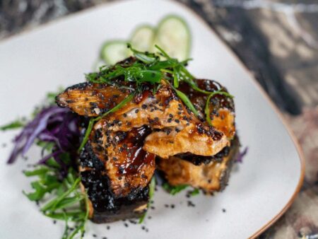 Glazed teriyaki fish with black sesame seeds, greens, and cucumber on white plate