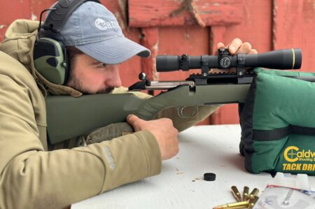 Man aims bolt-action rifle with scope at outdoor shooting range using rest bag.