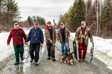 March of the Snowshoes: 4 Days of the Craziest Rabbit Hunting in the World Five hunters with dogs, guns, and snowshoes walking on a snowy rural road.