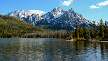 Snow-capped Sawtooth Mountains reflected in a calm alpine lake surrounded by evergreen forest.