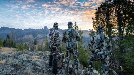 Three camouflaged hunters survey a scenic mountain wilderness at sunrise with bows