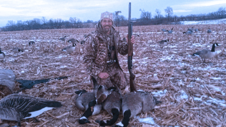 The 10 Gauge Hunter in camouflage kneeling with harvested Canada geese and shotgun in field.