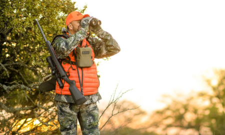 The Best Prime Day Hunting Deals on Amazon Right Now—Including a Ton of Sitka on Sale Hunter in blaze orange vest uses binoculars while carrying rifle in woods.