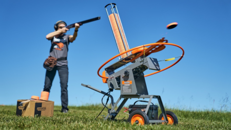 Person shooting skeet with orange clay target launcher machine on grassy field