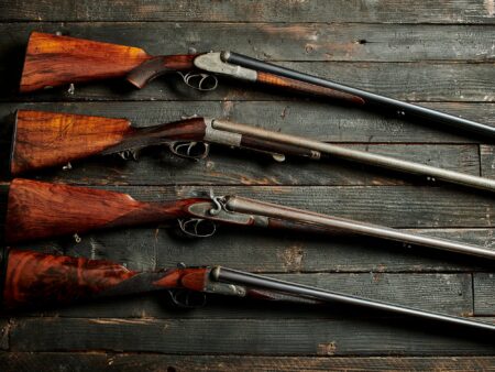 Double Take: Gorgeous Vintage Side-by-Side Shotguns That You Can Actually Afford Four vintage double-barrel shotguns with walnut stocks displayed on rustic dark wood surface.