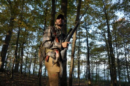 Hunter carrying rifle and harvested squirrel walking through autumn forest woodland