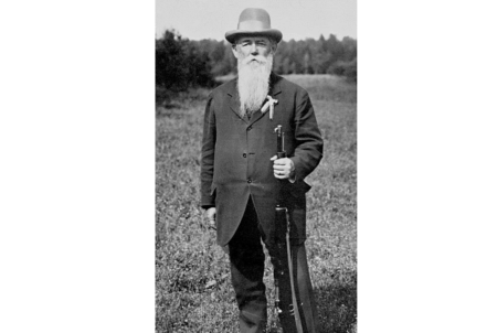 Elderly bearded man in Victorian-era suit and hat holding a walking cane outdoors.