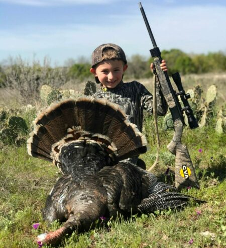 Young boy proudly poses with harvested wild turkey and CVA rifle outdoors.