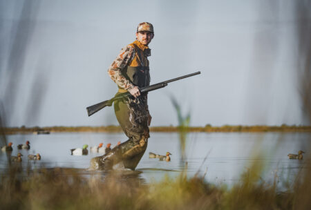Hunter in camouflage wading through marsh water carrying shotgun with duck decoys nearby