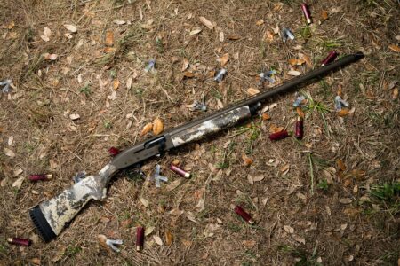 Camouflage shotgun lying on ground surrounded by spent red shotgun shells outdoors