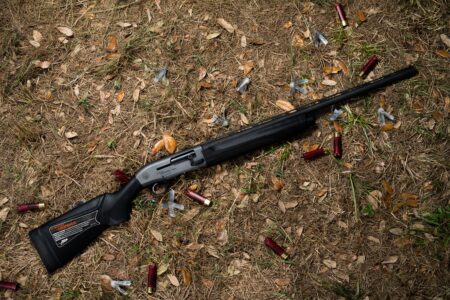 Black semi-automatic shotgun lying on ground surrounded by spent red shotgun shells
