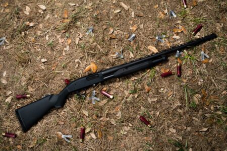 Black pump-action shotgun lying on ground surrounded by spent red shotgun shells.