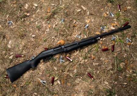 Black pump-action shotgun lying on ground surrounded by spent red shotgun shells.