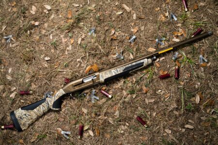 The Browning Maxus II: Tested and Reviewed Camouflage pump-action shotgun lying on ground surrounded by spent red shotgun shells.