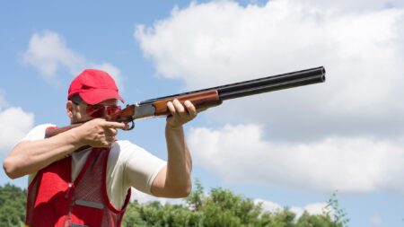 Man in red vest and cap aiming shotgun skyward during clay pigeon shooting