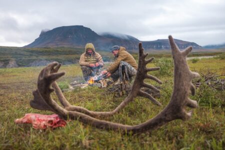 Two hunters sit by campfire in wilderness with caribou antlers in foreground.