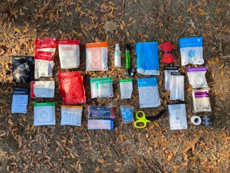 Outdoor first aid kit contents spread on ground including bandages, QuikClot, and medical supplies