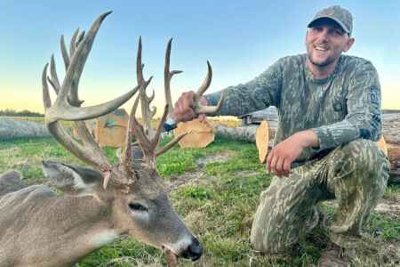 Smiling hunter in camouflage posing with large trophy whitetail buck outdoors