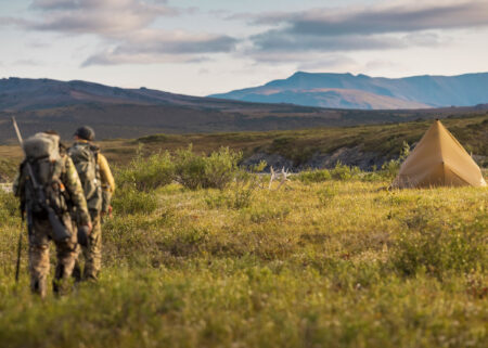 Two camouflaged hunters with backpacks approach a canvas tipi tent on Arctic tundra.